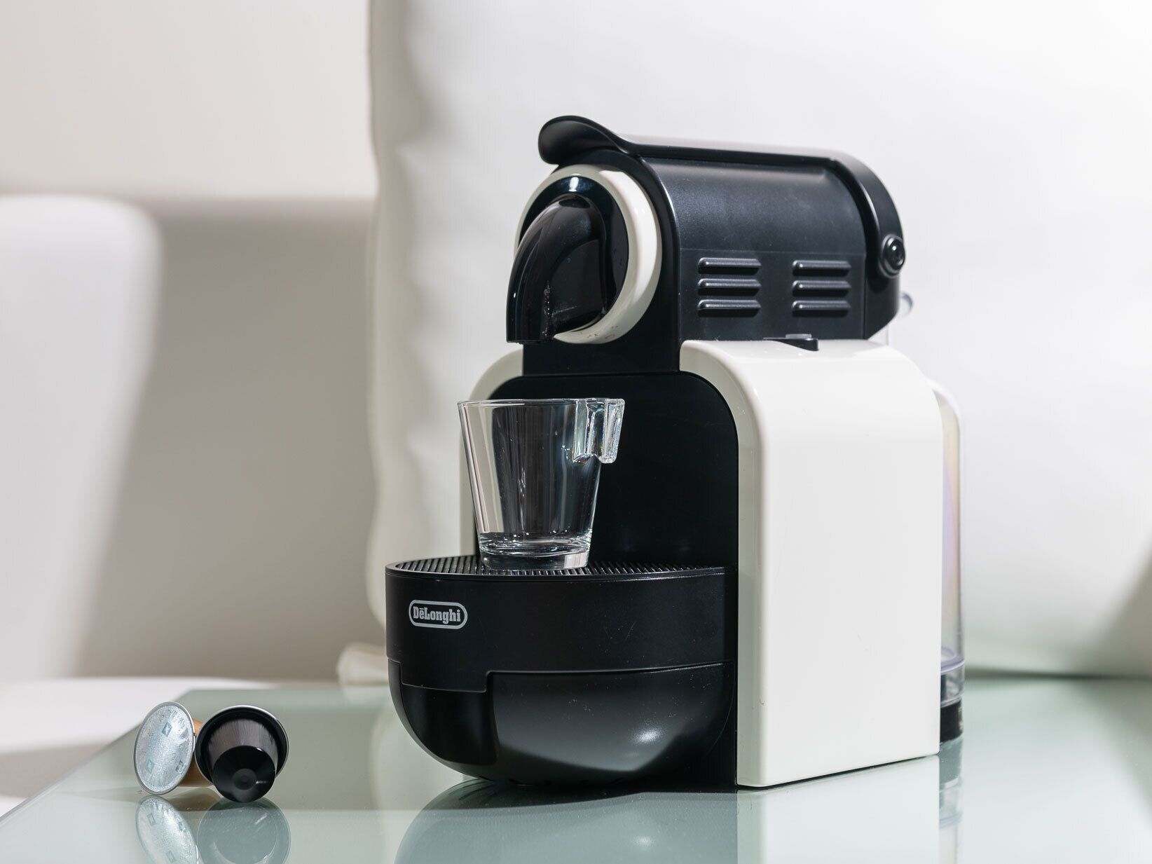 Uma máquina de café Delonghi com um suporte de vidro para chávenas numa superfície refletora.
