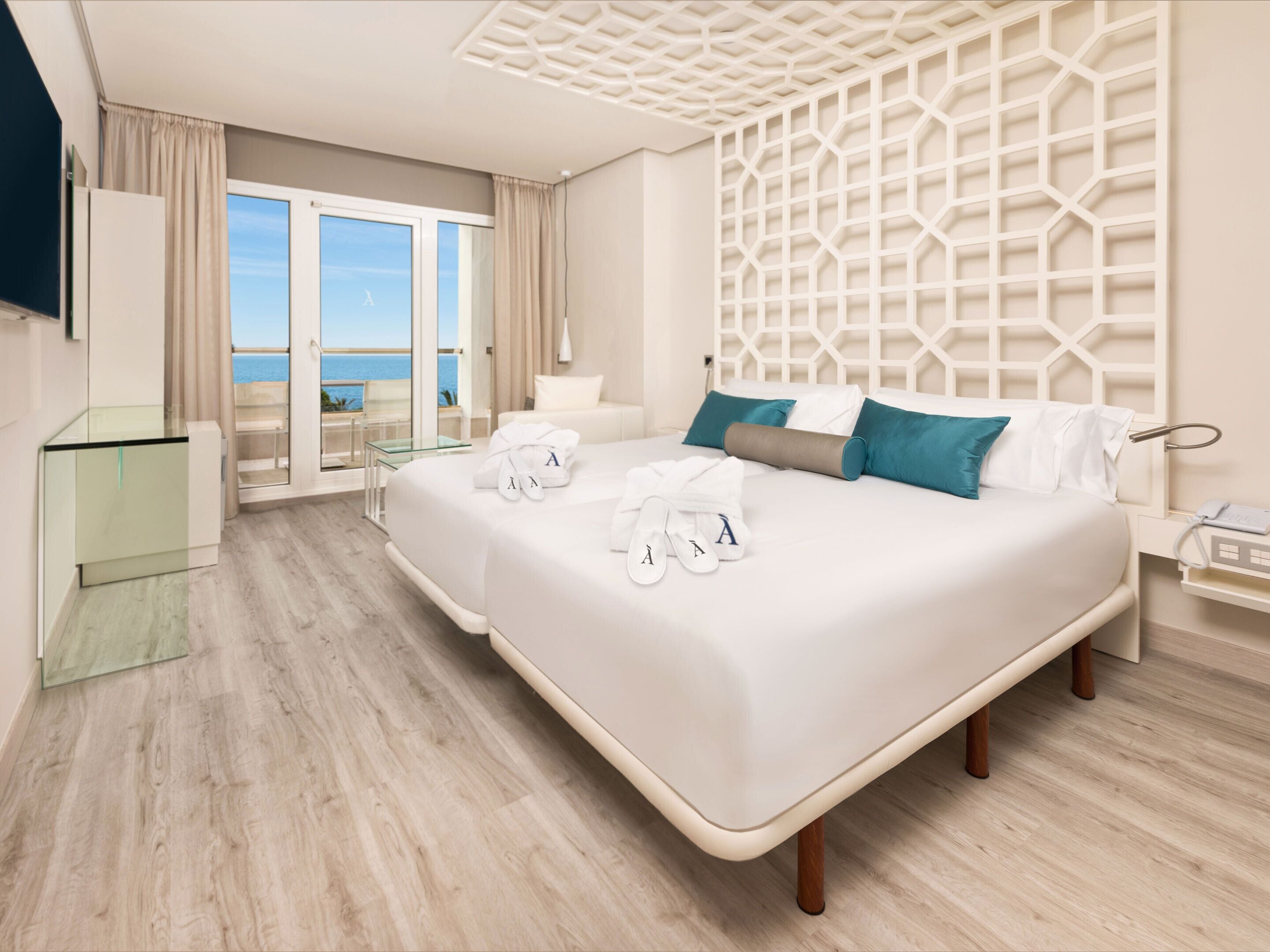 A modern hotel room with a large bed, a glass desk, a view of the sea, and a decorative wall panel.