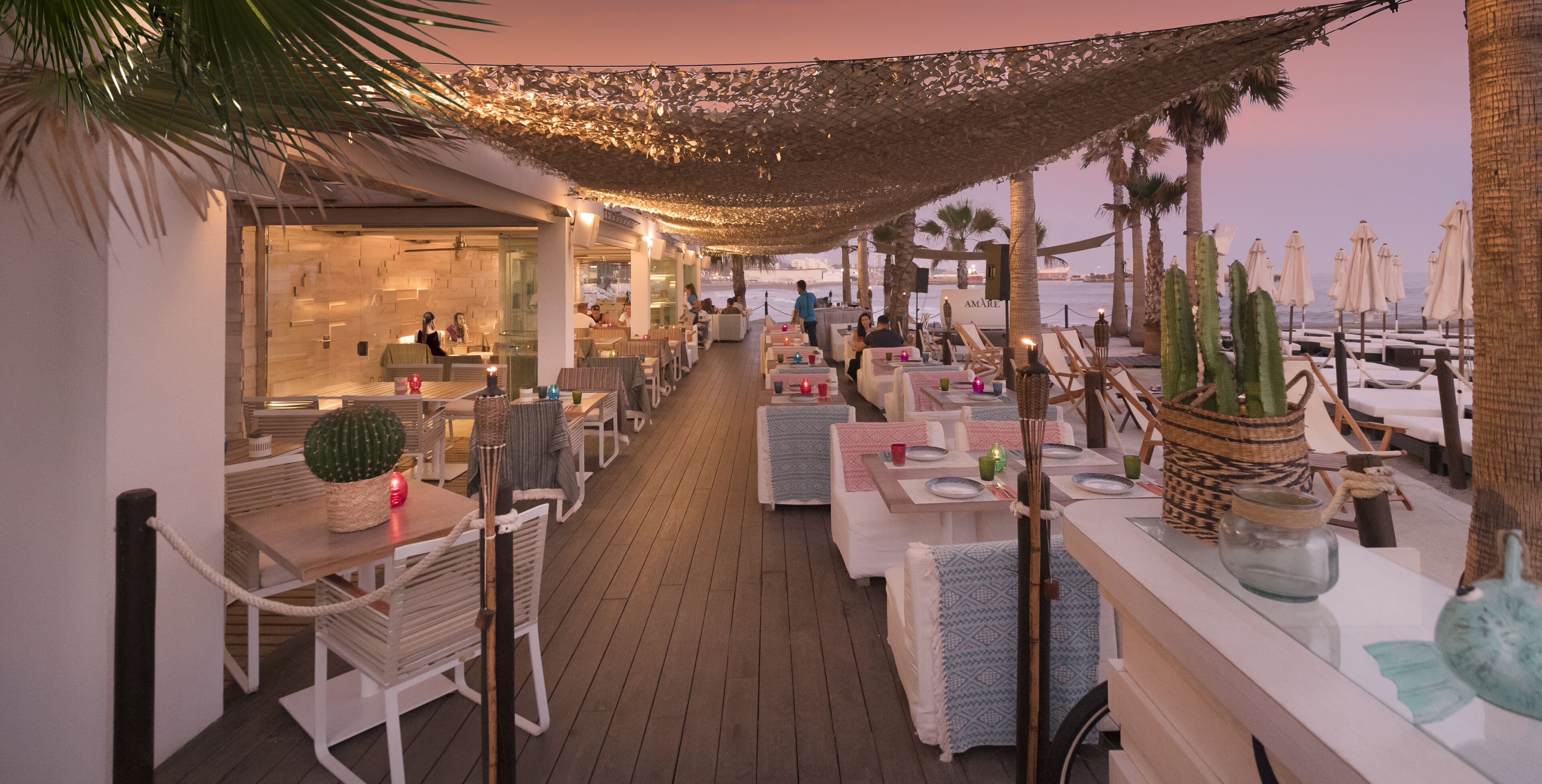 Un espacio exterior para comer con mesas y sillas, palmeras y vistas al mar.