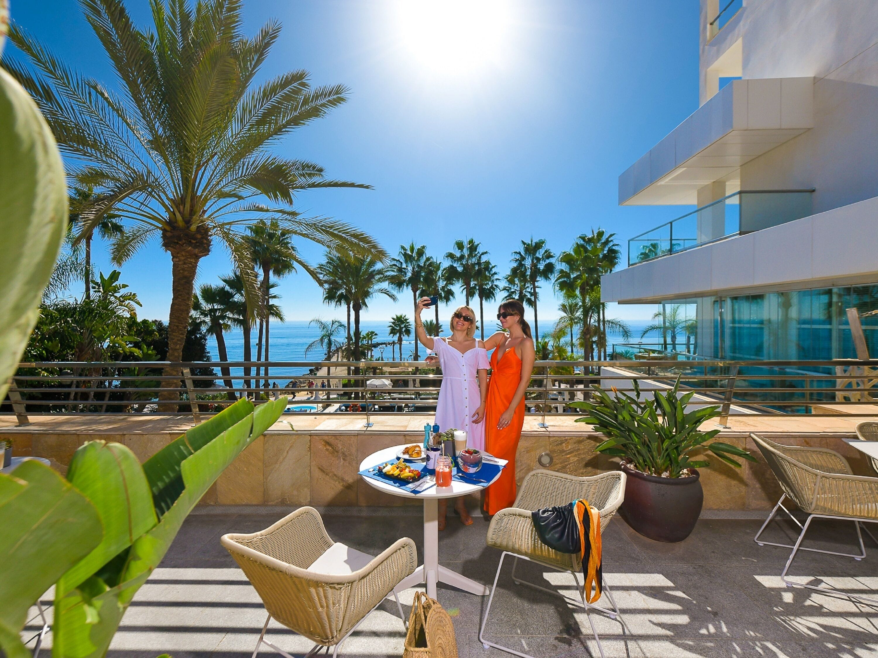 A rooftop terrace with palm trees, a pool, and a dining area with wicker chairs and a table.