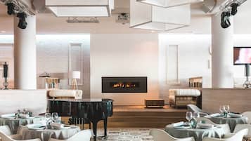 Un hall d’hôtel moderne, doté d’un grand piano, d’une cheminée et d’un espace repas avec des tables et des chaises.