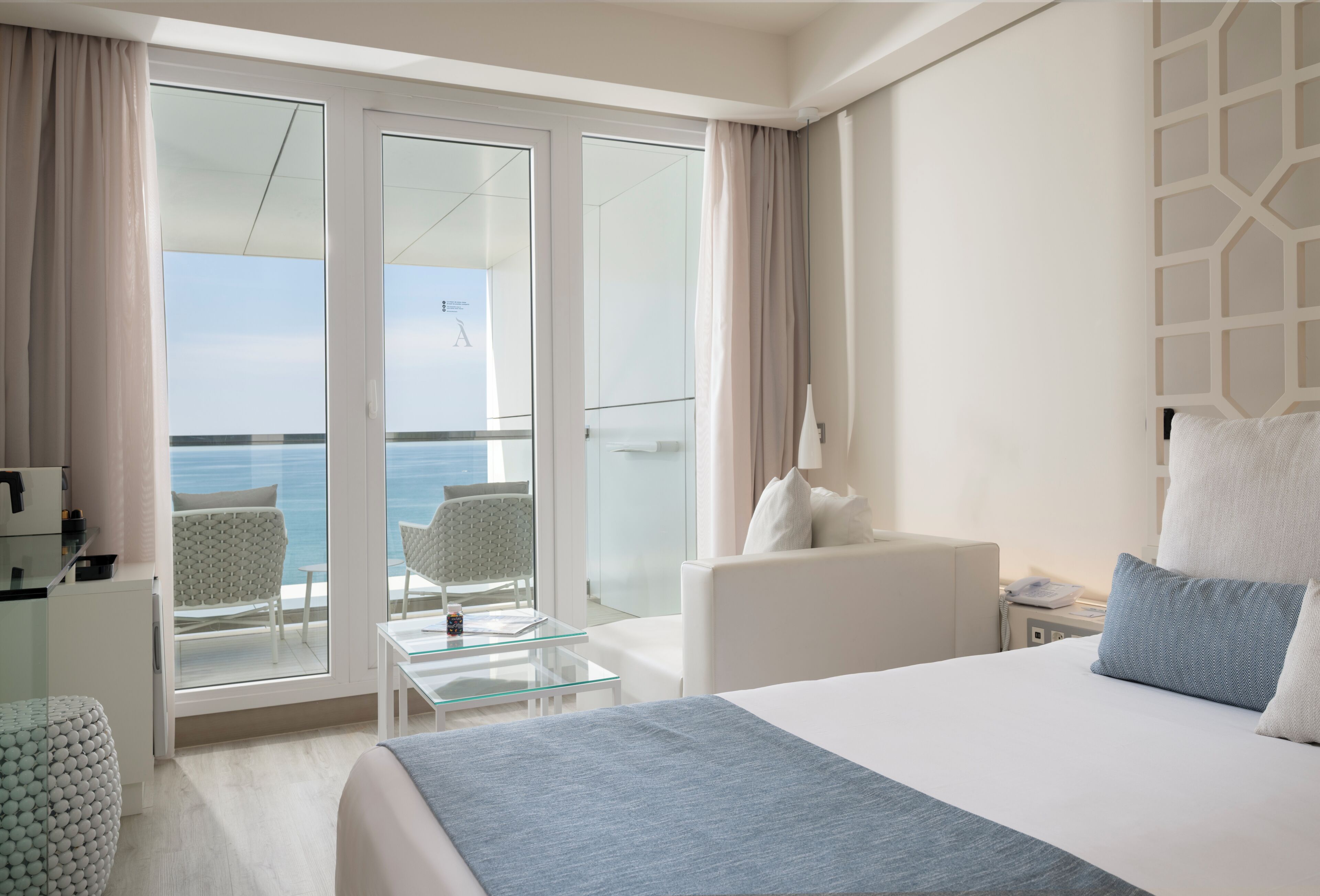 A hotel room with a bed, a glass table, a chair, and a view of the sea through large glass doors.