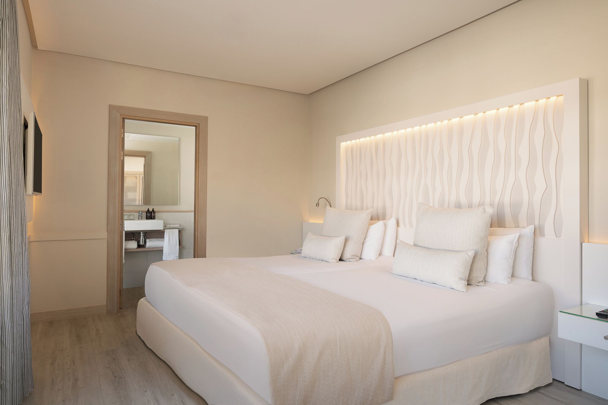 Moderni hotellihuone, jossa on suuri sänky, yöpöytä, peili ja kylpyhuone, joka näkyy avoimesta ovesta.
