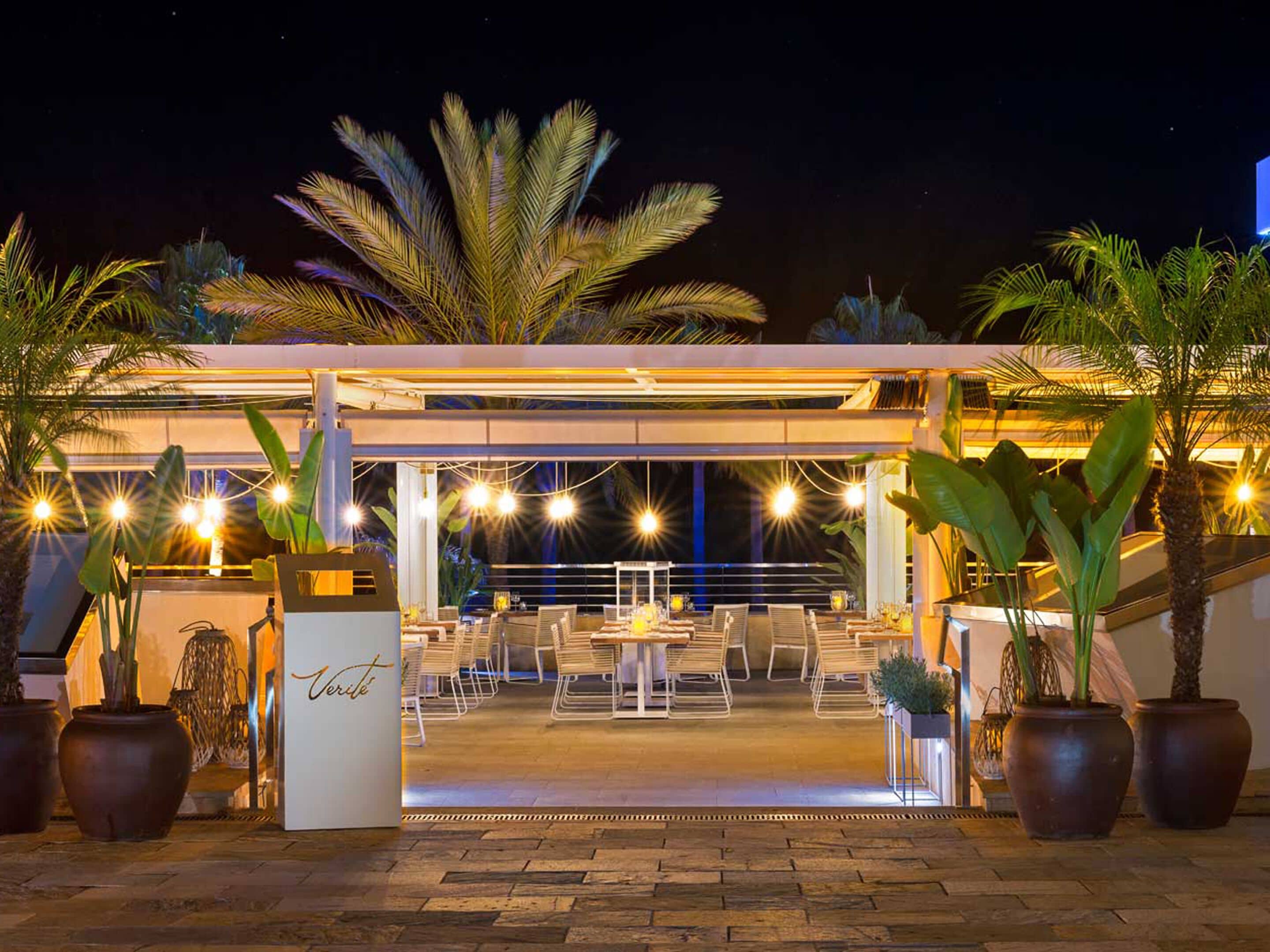 An outdoor dining area with tables and chairs, surrounded by potted plants and palm trees, under a covered structure with string lights.