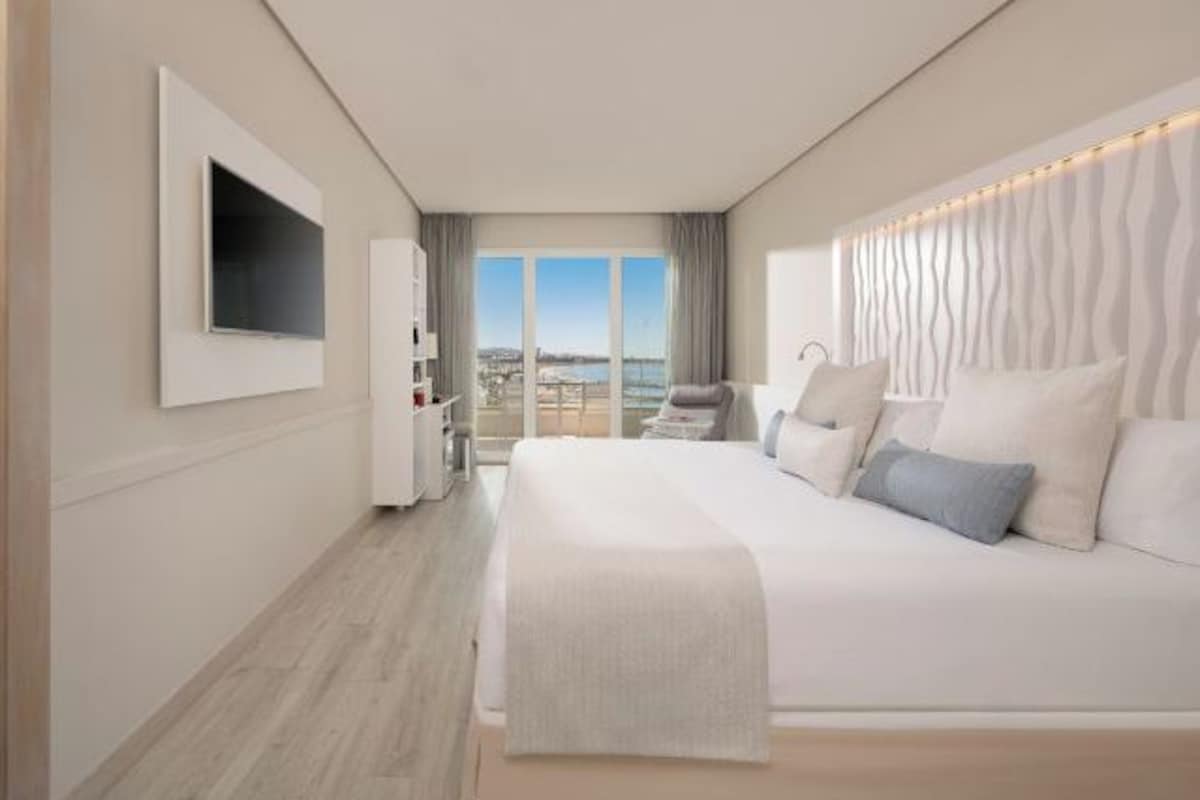 A modern hotel room with a large bed, a TV mounted on the wall, a balcony with a view, and a decorative headboard.