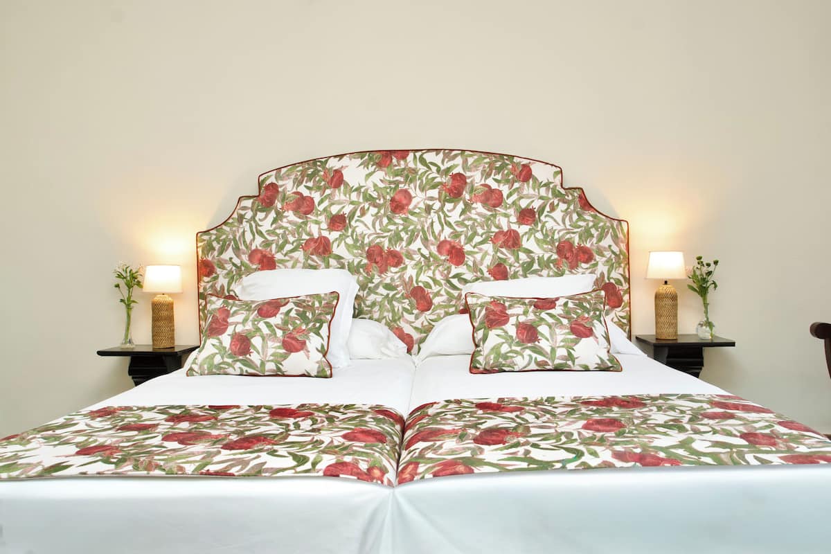 Cama doble con ropa de cama y cabecera estampada, dos mesitas de noche con lámparas y un florero con planta en cada una.