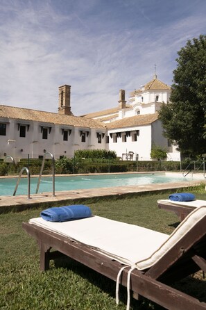 Zona de piscina con sillas de descanso y un edificio blanco de fondo con tejado de azulejos.