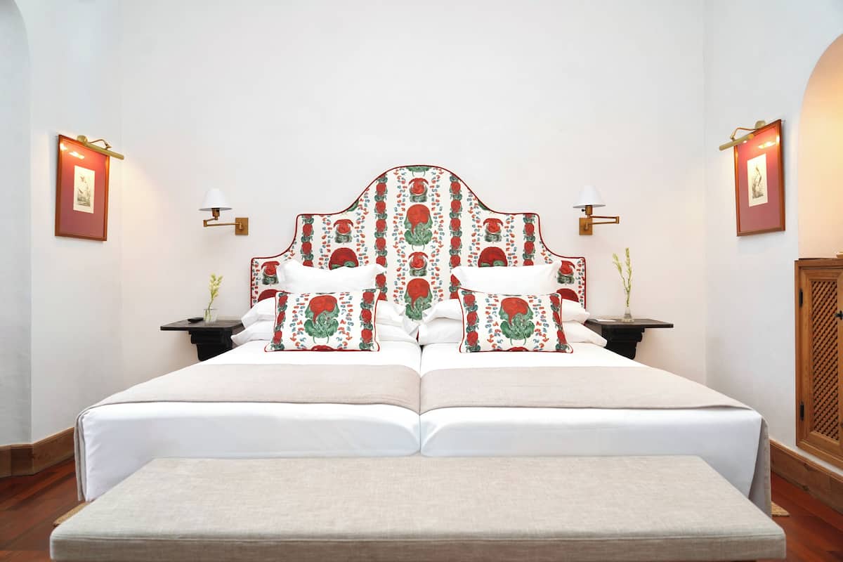 Una cama doble con cabecera floral, dos mesitas de noche con lámparas y un banco al pie de la cama.
