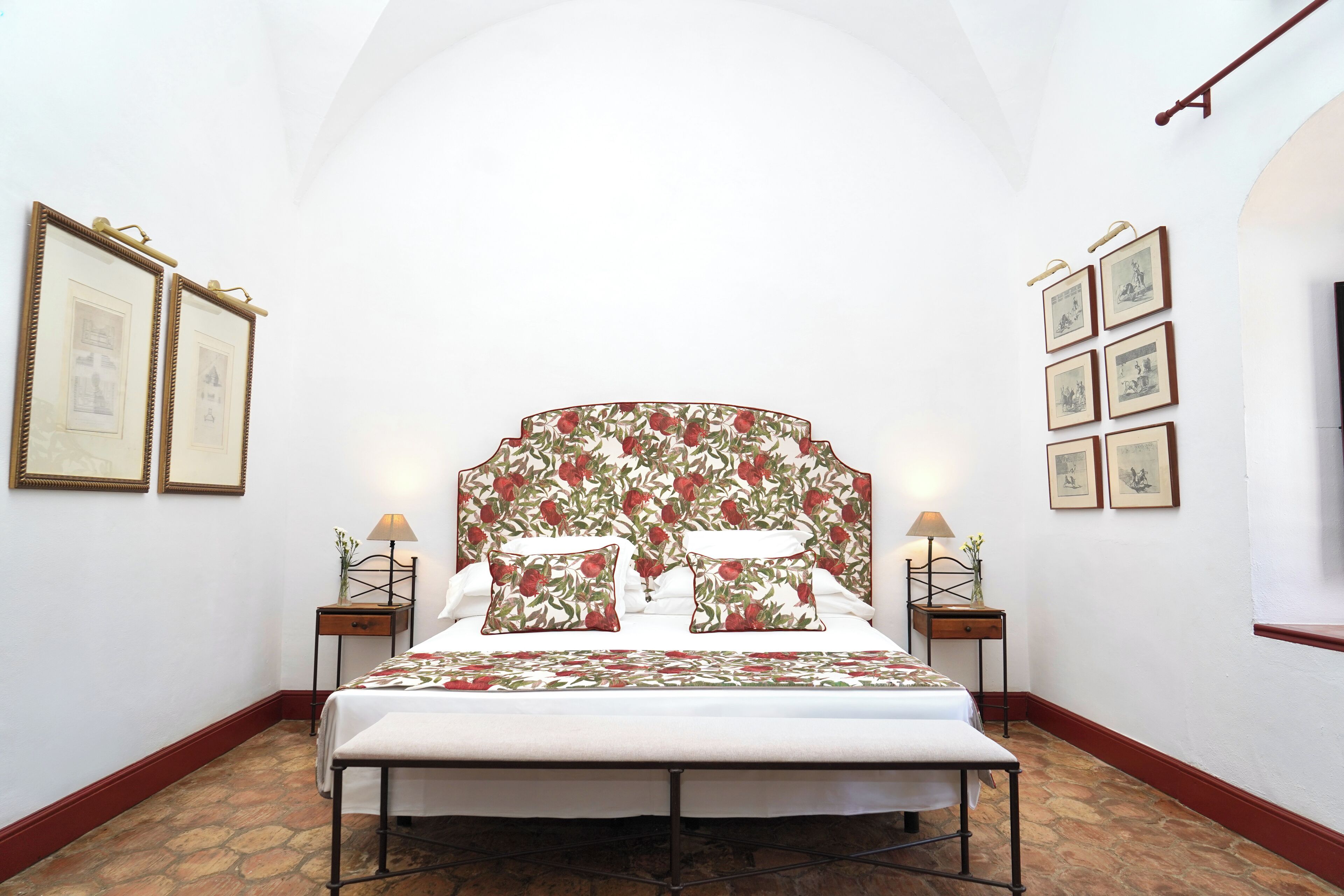 Un dormitorio con una cama grande, cabecera de flores, dos mesitas de noche con lámparas y cuadros enmarcados en las paredes.