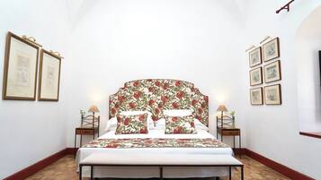 Un dormitorio con una cama grande, cabecera de flores, dos mesitas de noche con lámparas y cuadros enmarcados en las paredes.