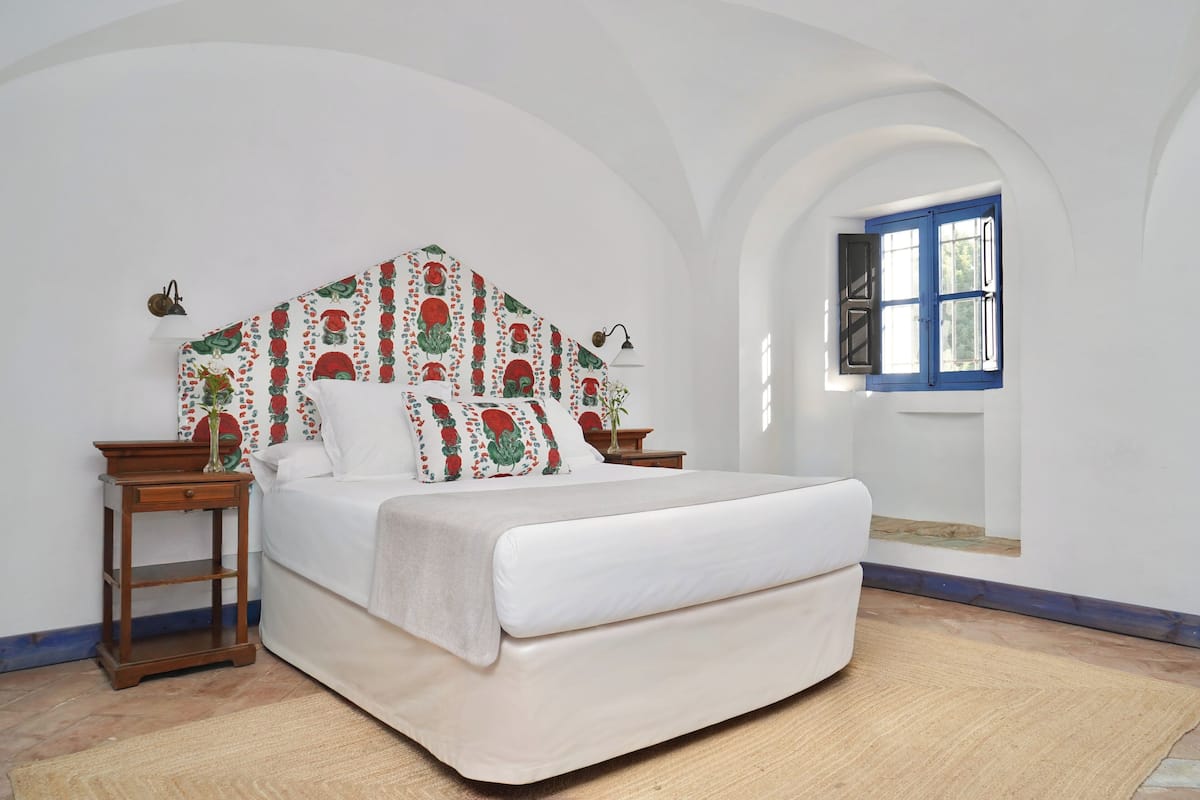 Un dormitorio con cama, dos mesitas de noche, piso de madera, pared blanca, ventana con contraventanas azules y cabecero estampado.