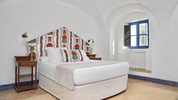 Een slaapkamer met een bed, twee nachtkastjes, een houten vloer, een witte muur, een raam met blauwe luiken, en een gestreept hoofdeinde.