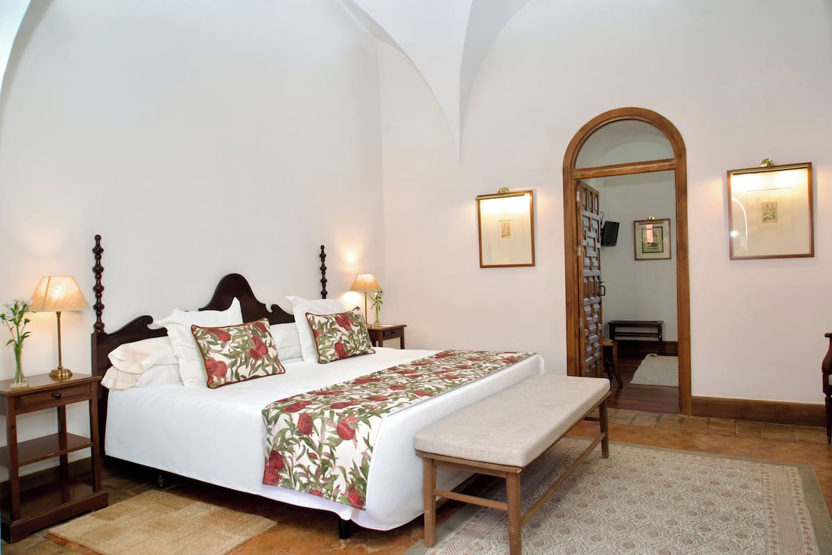 Un dormitorio con una cama grande, mesitas de noche, un banco, un espejo y cuadros en la pared.