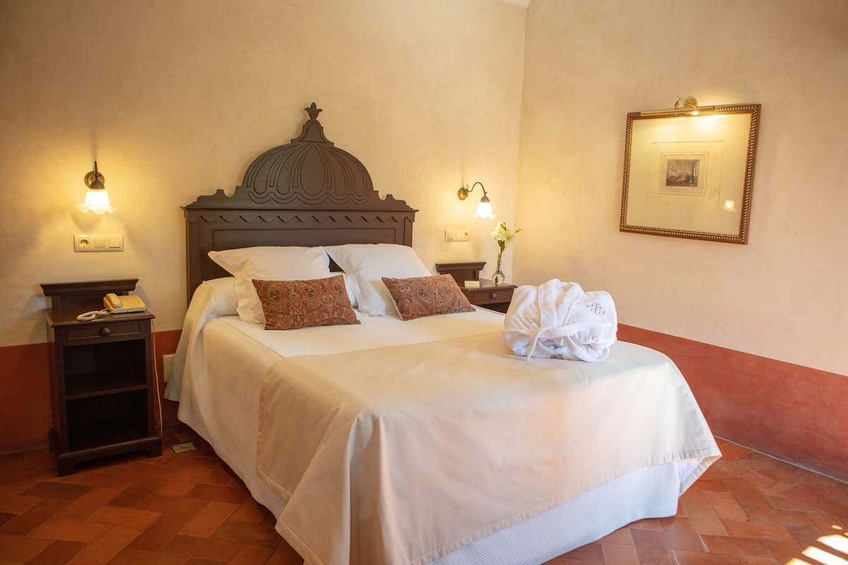 Habitación de hotel con una cama grande, dos mesitas de noche, un buró y un cuadro colgado en la pared, además de una lámpara de pared.