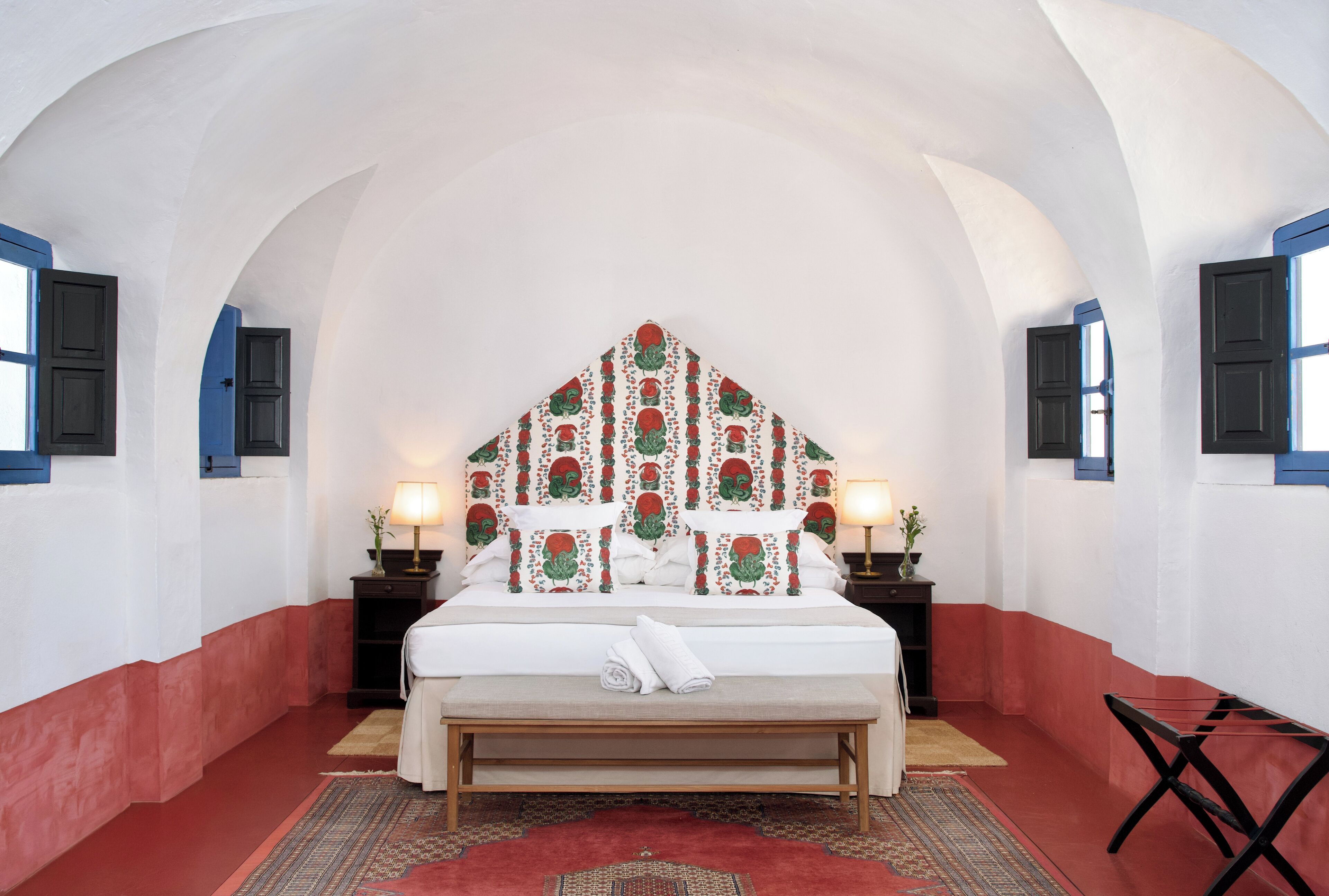 Un dormitorio con una decoración en blanco y rojo, una cama grande con cabecera estampada, dos mesitas de noche con lámparas, y un banco al pie de la cama.