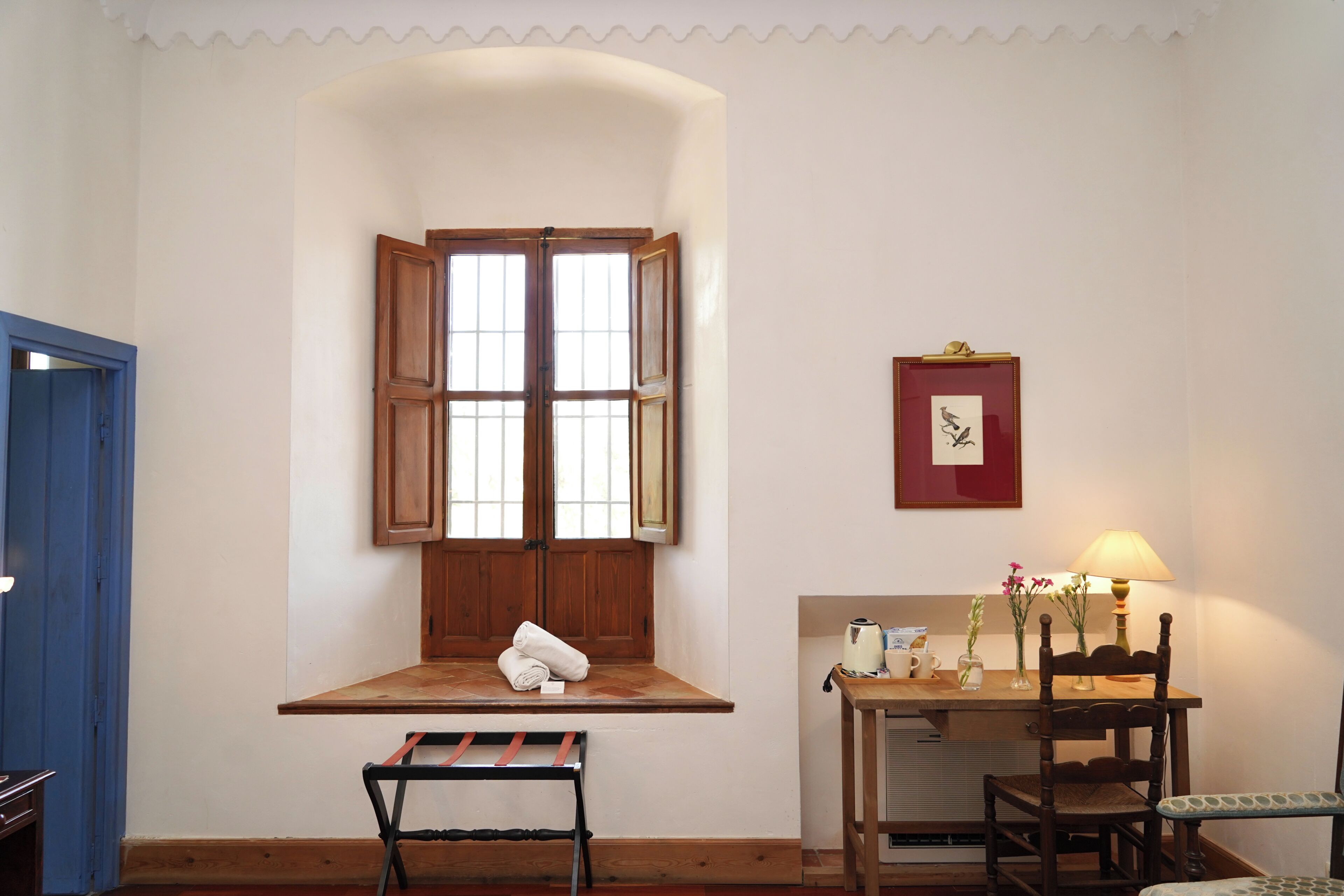Una habitación con ventana, una mesa con lámpara, una silla y un banco.
