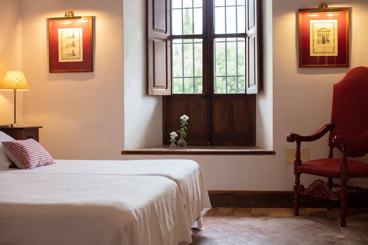 Habitación de hotel con una cama, una silla, una ventana con contraventanas abiertas y cuadros en las paredes.