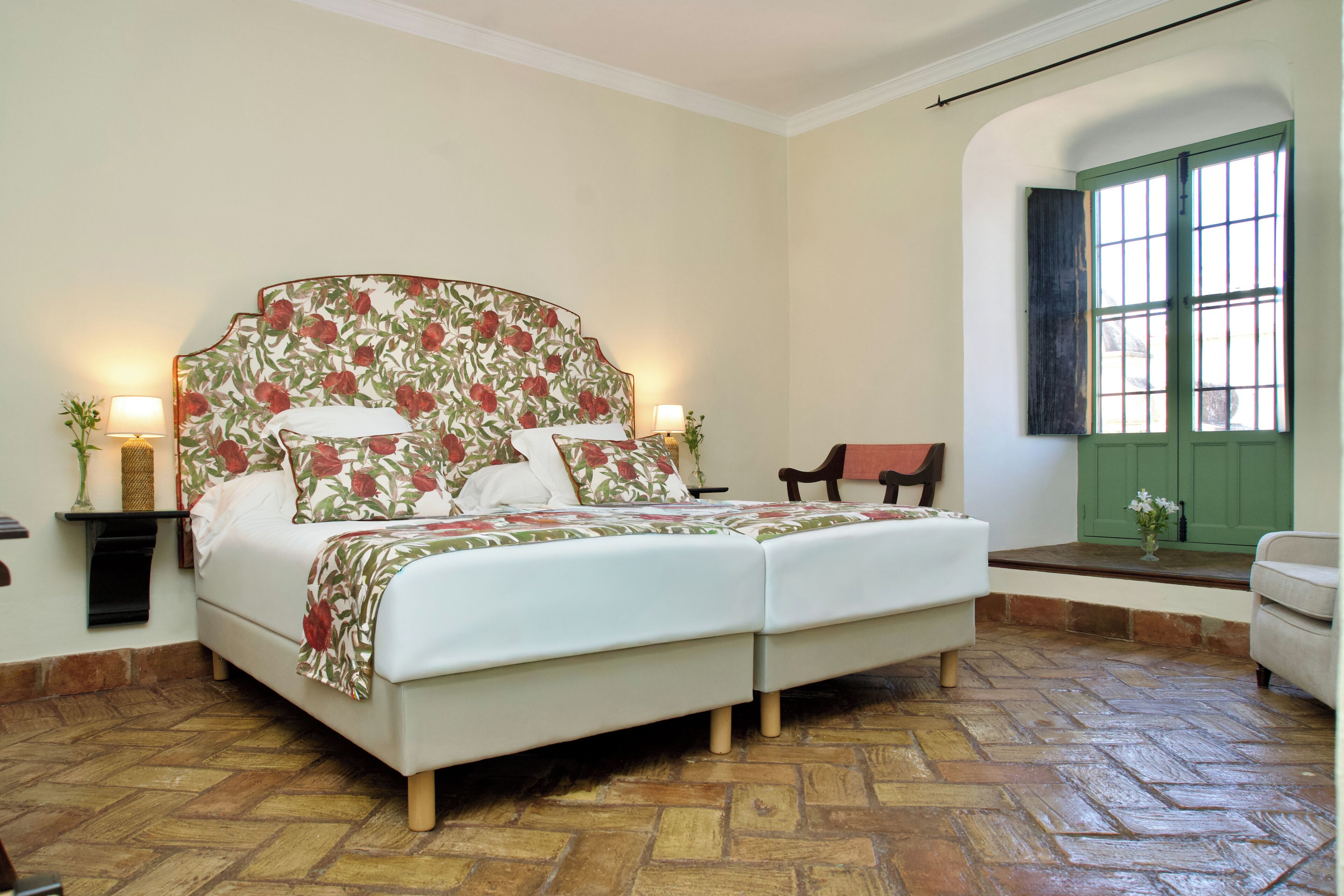 Una cama doble con cabecera floral, dos lámparas de noche y una puerta verde al fondo.