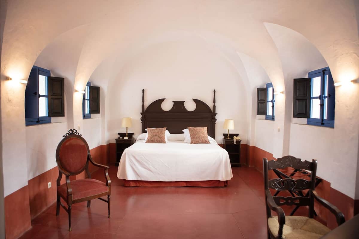 Un dormitorio con una cama grande, dos mesitas de noche, una silla y una ventana con contraventanas.