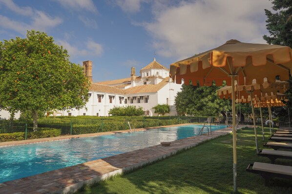 A swimming pool with a tiled deck, a white building with a dome roof, and a large umbrella structure. - Monasterio de San Francisco (Palma del Rio)
