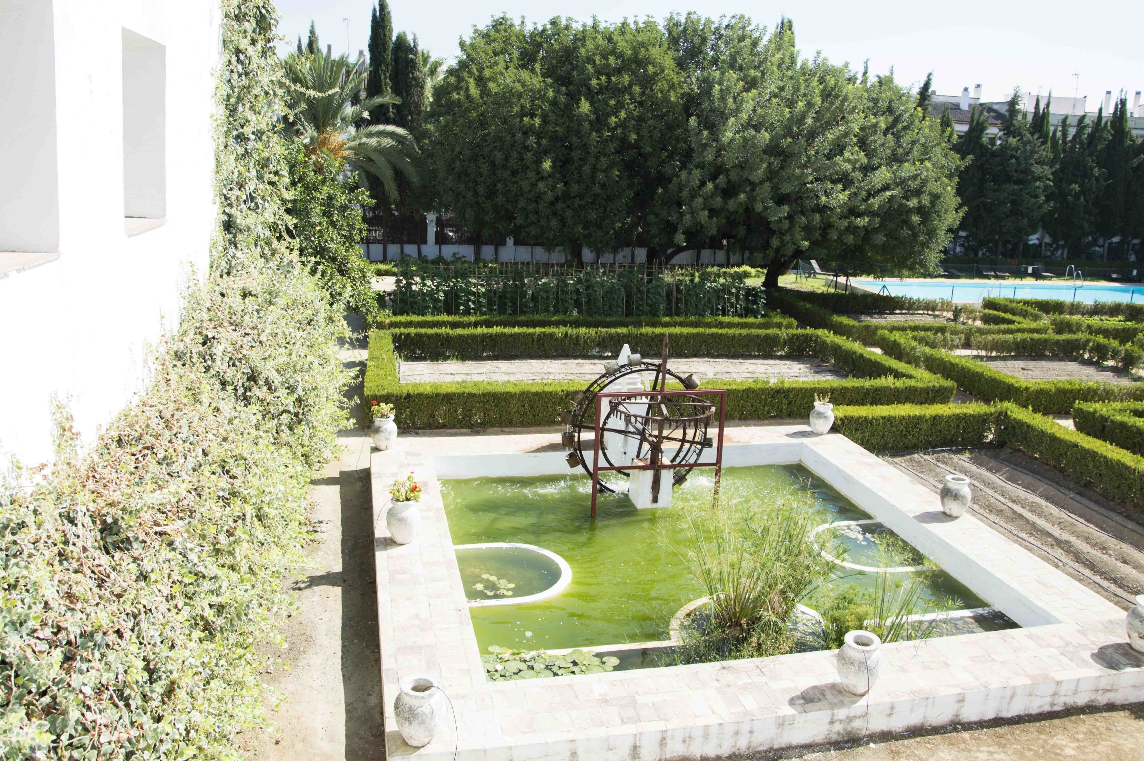 A formal garden with a central fountain, neatly trimmed hedges, and a pool in the background.