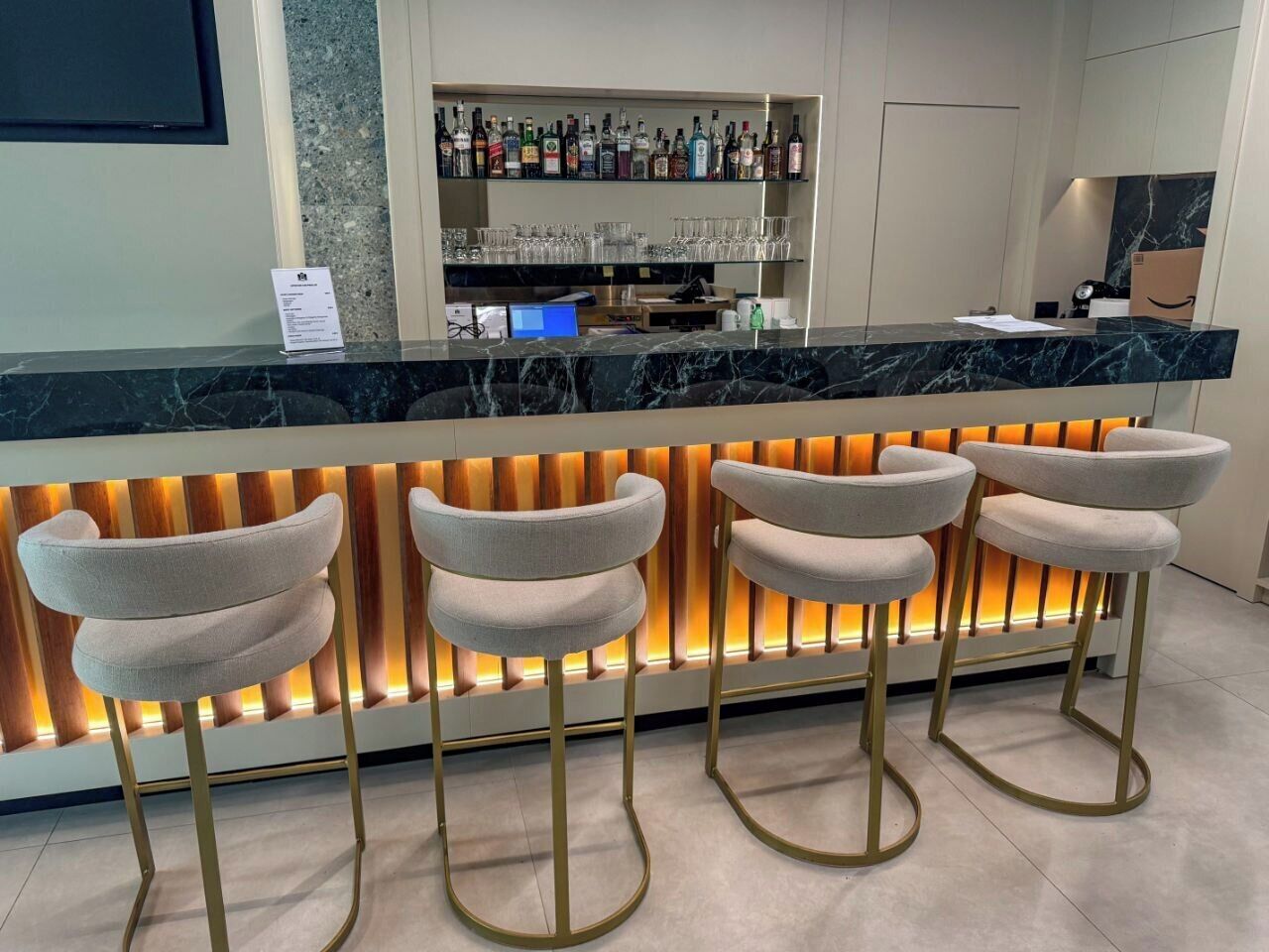 A bar area with a marble countertop, four upholstered bar stools, a shelf with liquor bottles, and a lit-up wall panel.