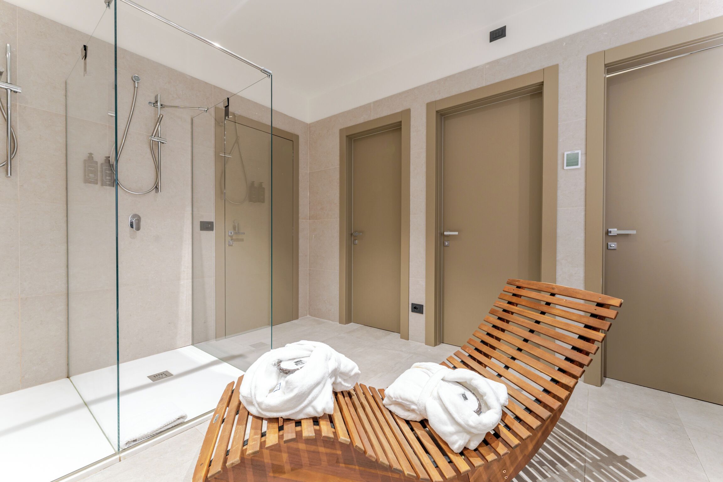 A modern bathroom with a glass shower enclosure, wooden bench, and two towels.