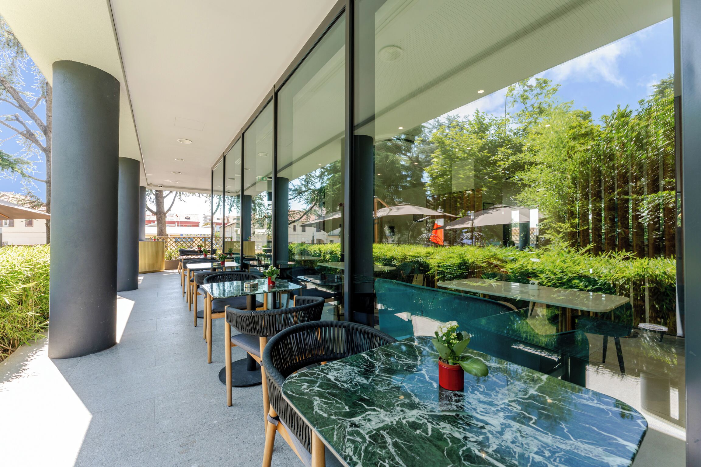 Un espace extérieur aménagé avec des tables et des chaises, entouré de murs et de piliers en verre, donnant sur un jardin.