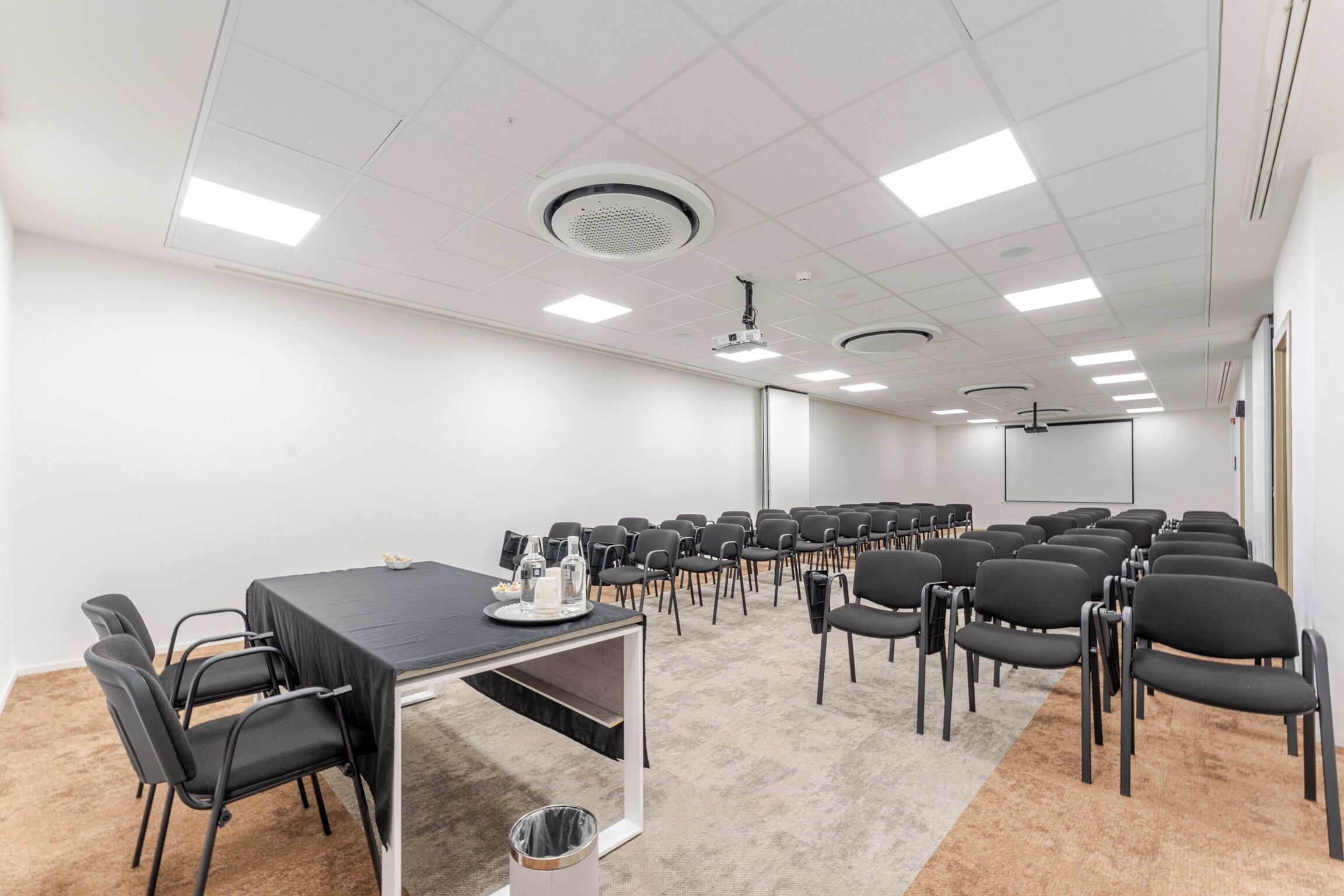 A conference room with a long table, chairs, carpeted floor, white walls, ceiling lights, and a projector.