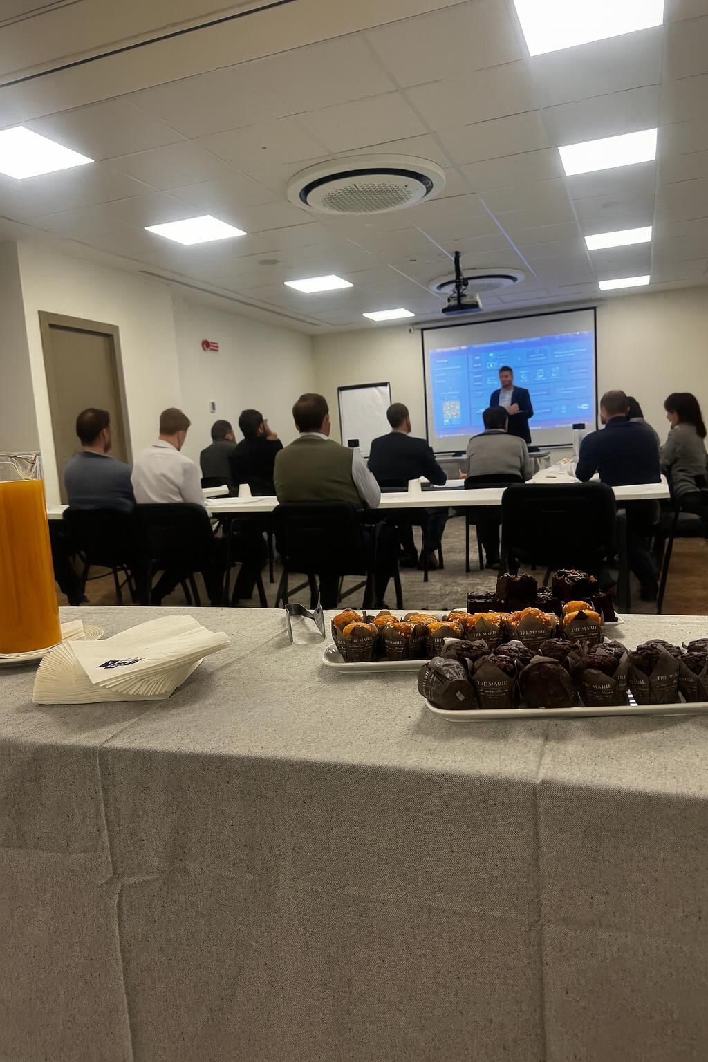 A conference room with a presentation screen, attendees seated at tables, and a table with pastries.