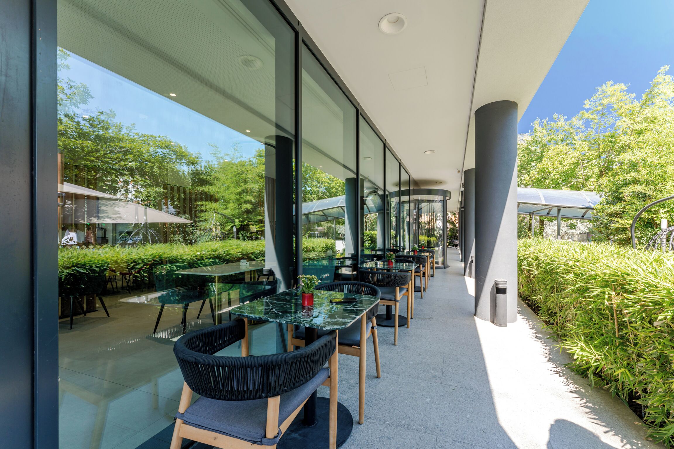An outdoor dining area with glass tables and chairs, surrounded by greenery and a clear sky.