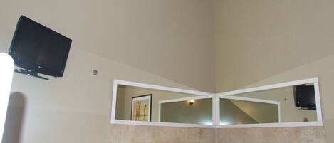 A bathroom with a bathtub, a ceiling fan, a TV mounted on the wall, and a mirror above the bathtub.
