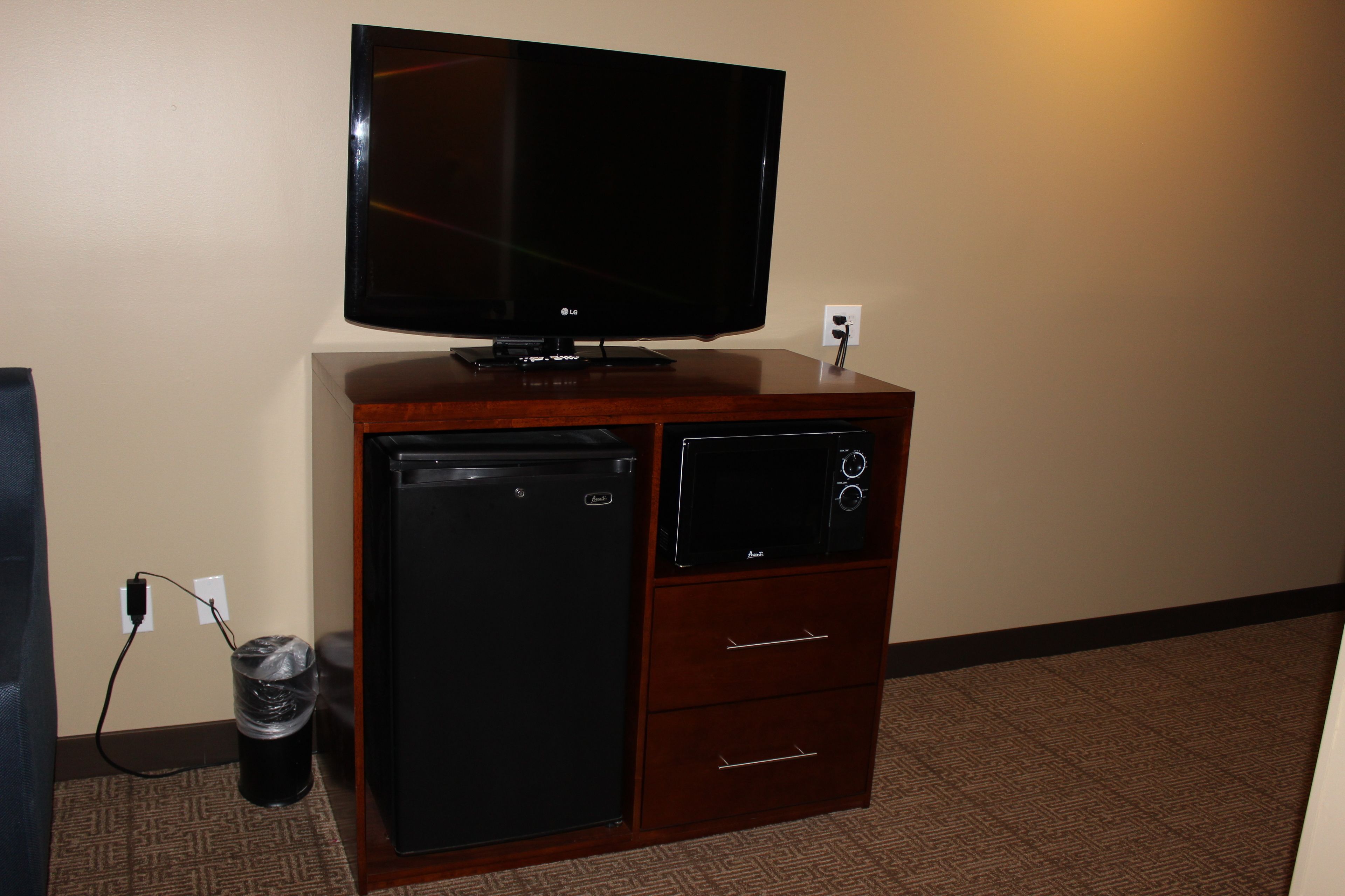 A hotel room with a flat-screen TV on a wooden cabinet, a mini-fridge, a small trash can, and a power outlet.