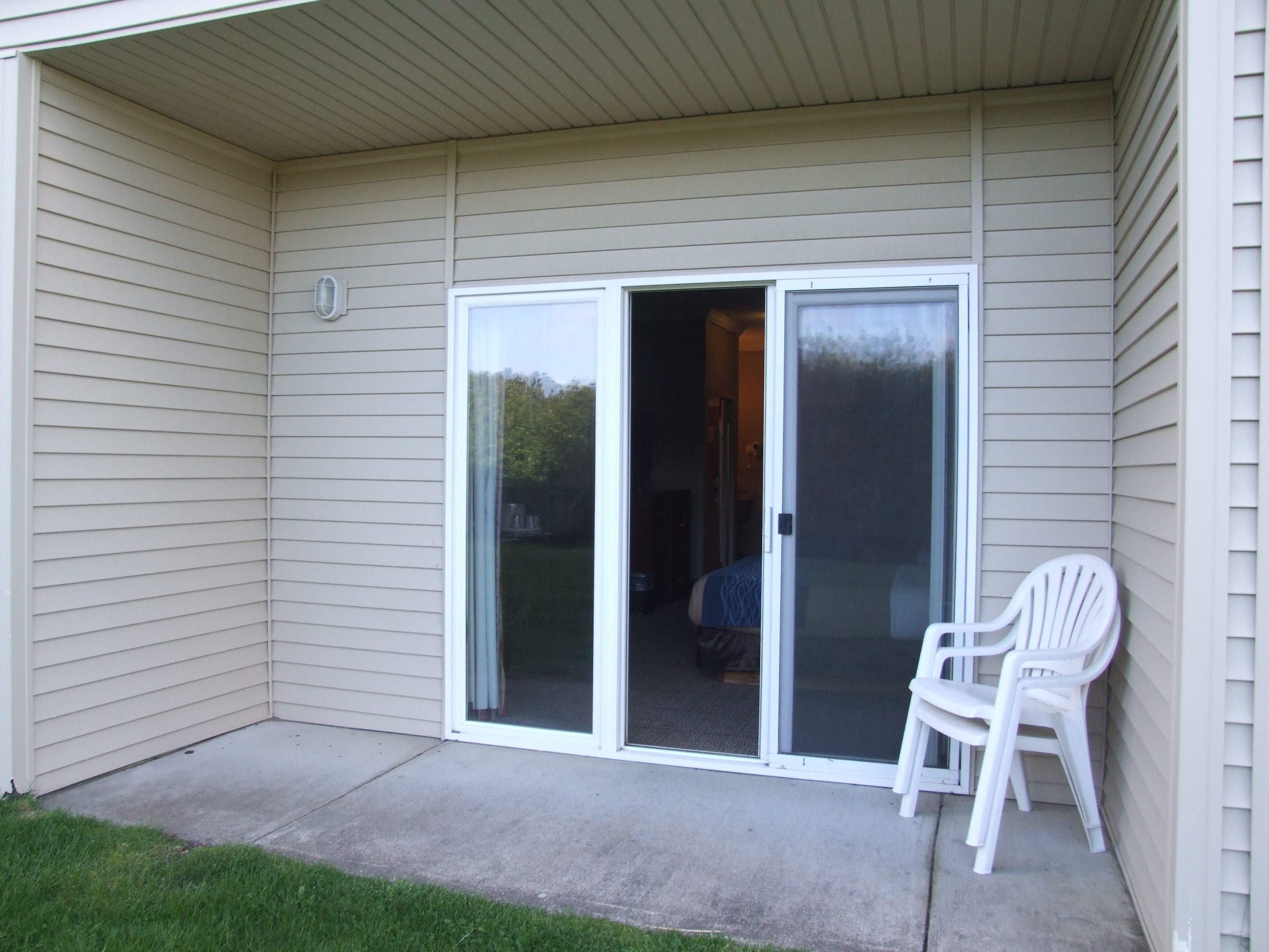 A covered patio with a sliding glass door leading to a bedroom. There is a white plastic chair on the patio.