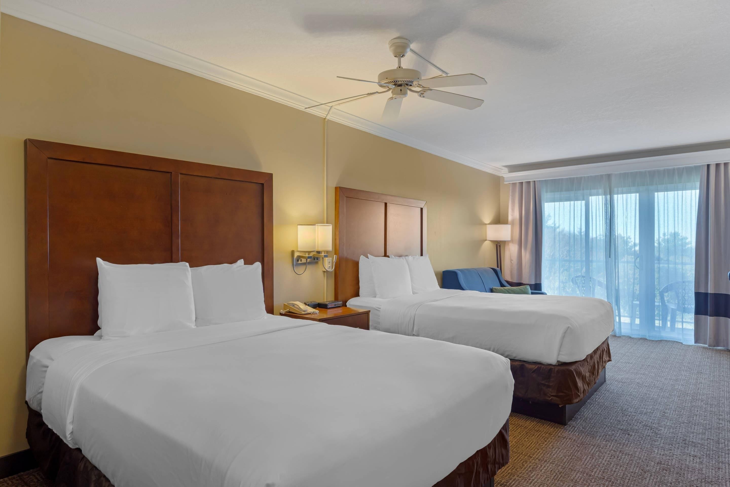 A hotel room with two beds, a ceiling fan, a nightstand with a phone, and a view of the outdoors through large windows.