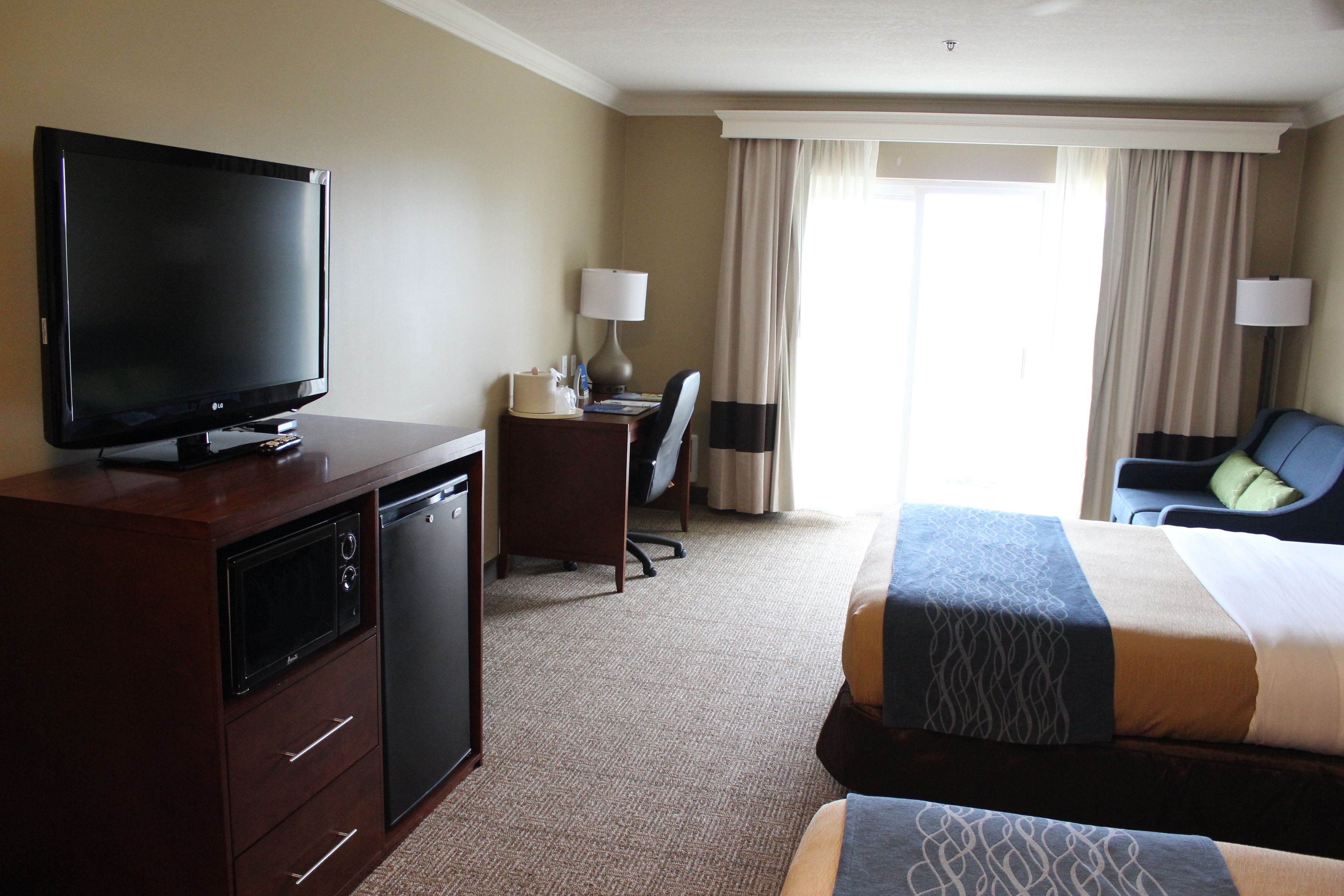 A hotel room with a flat-screen TV, a desk with a lamp, a chair, a bed with patterned bedding, and a view of the outdoors through the window.