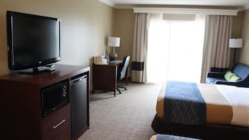 A hotel room with a flat-screen TV, a desk with a lamp, a chair, a bed with patterned bedding, and a view of the outdoors through the window.