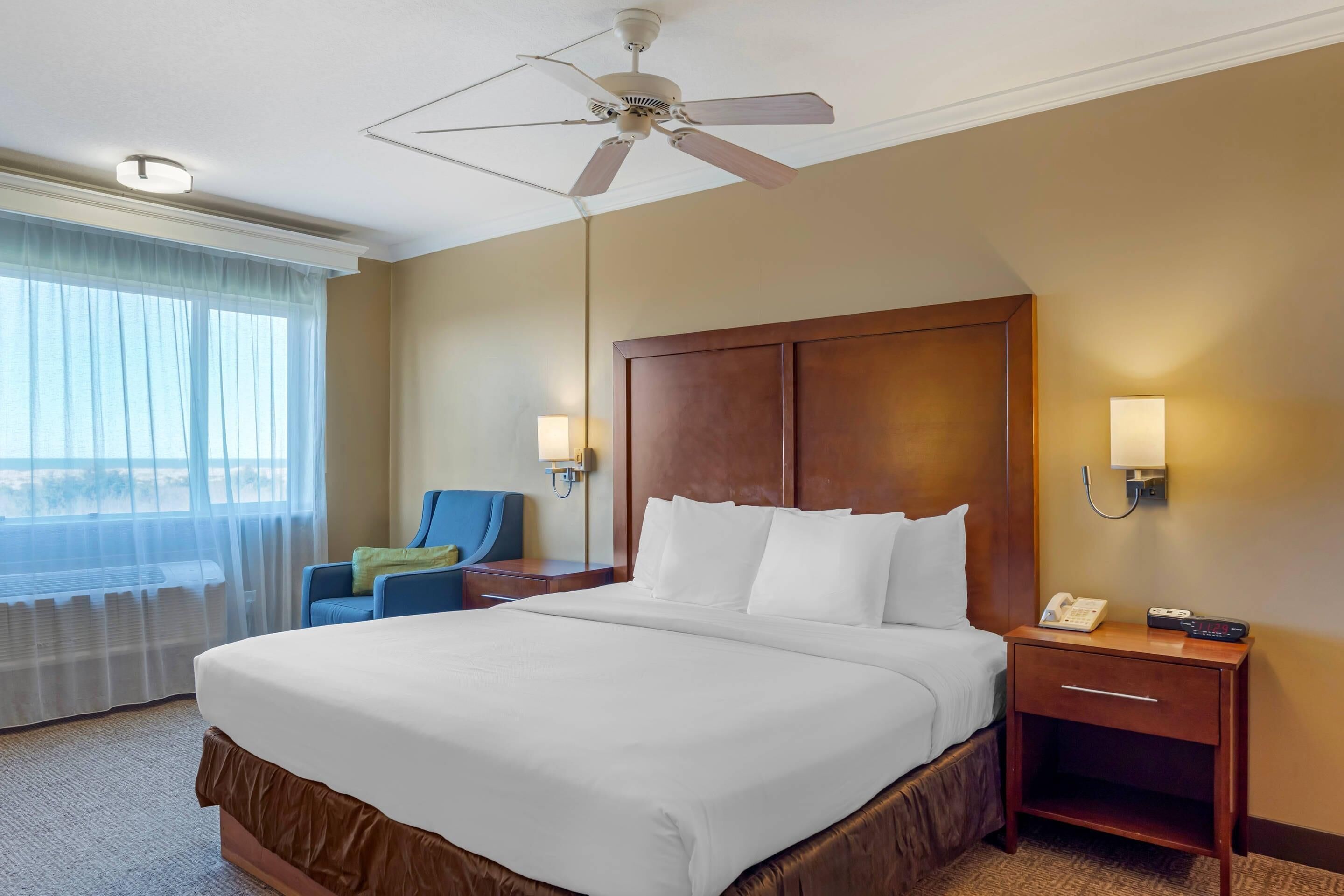 A hotel room with a large bed, a ceiling fan, a nightstand with a phone and remote, a chair, and a window with sheer curtains.