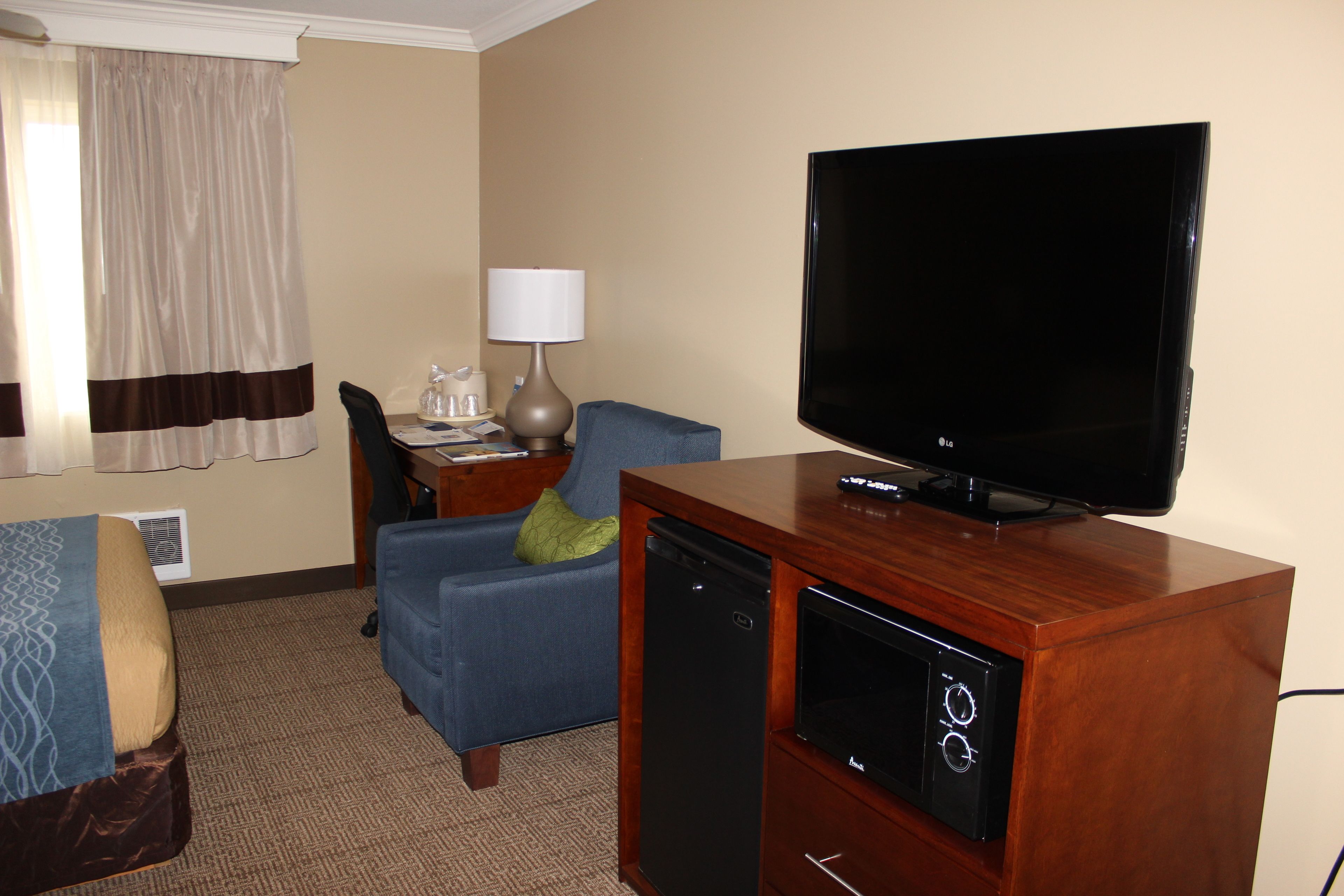 Habitación de hotel con televisión sobre un soporte de madera, un sillón azul, un pequeño escritorio con lámpara y una cama con manta estampada.