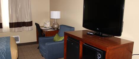 Habitación de hotel con televisión sobre un soporte de madera, un sillón azul, un pequeño escritorio con lámpara y una cama con manta estampada.
