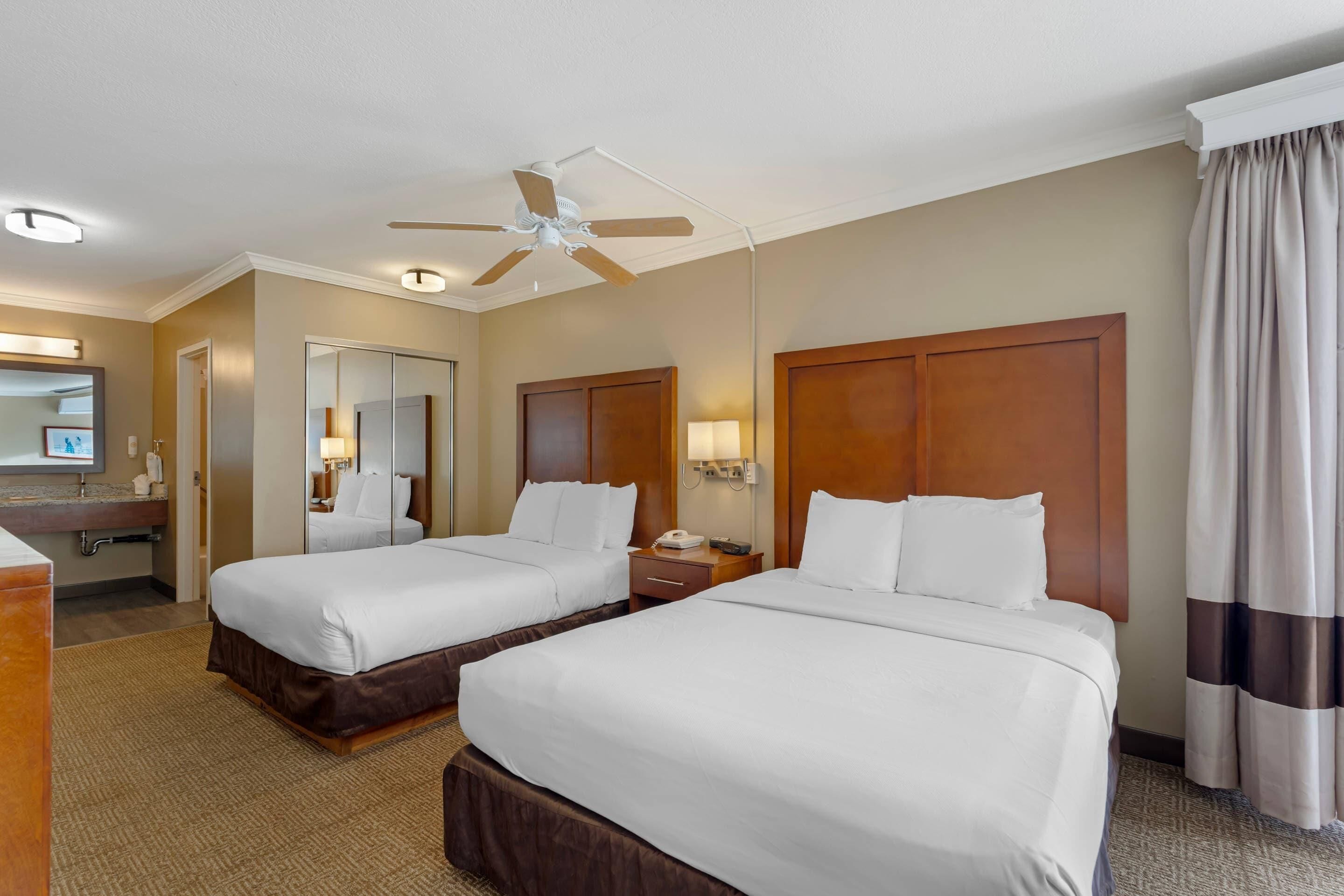 Une chambre d’hôtel avec deux lits, un ventilateur de plafond, un miroir, une coiffeuse avec un lavabo et une fenêtre avec des rideaux.