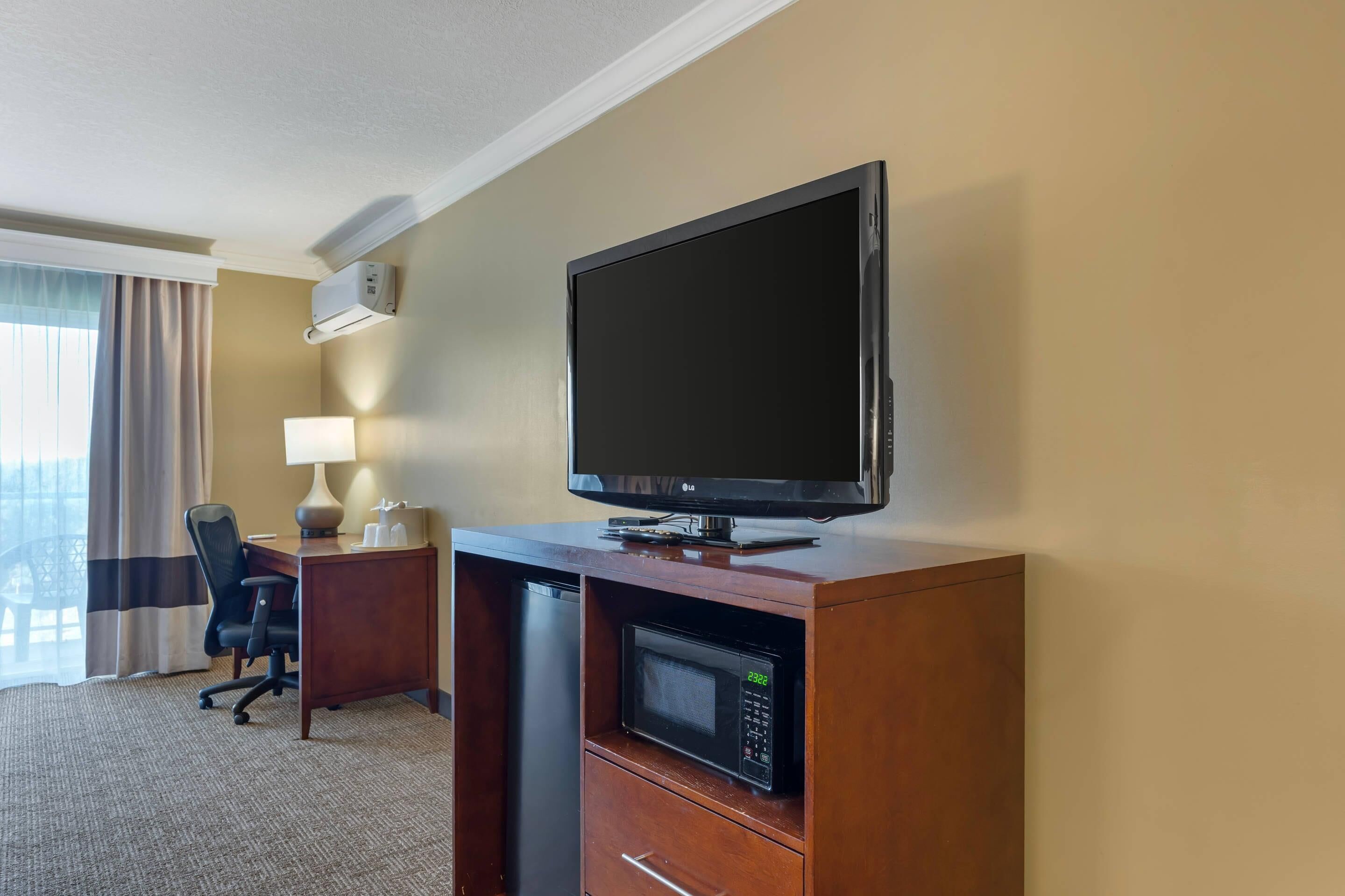 A hotel room with a flat-screen TV, a microwave, a desk with a chair, and a lamp.