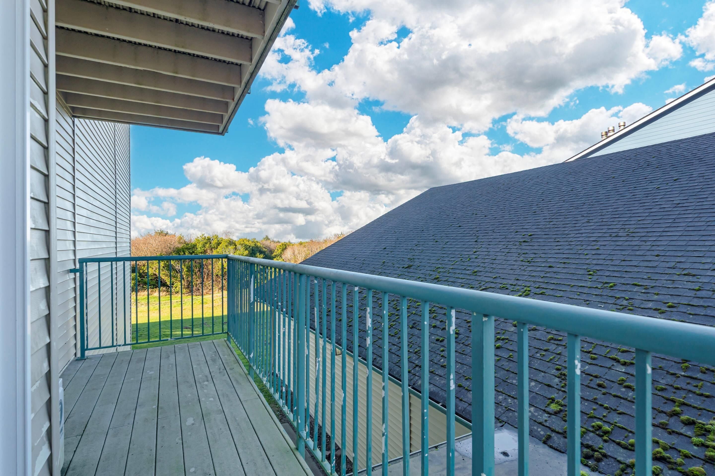 Une terrasse en bois avec une rambarde de couleur turquoise, un ciel dégagé parsemé de nuages et un bâtiment doté d’un toit sombre.