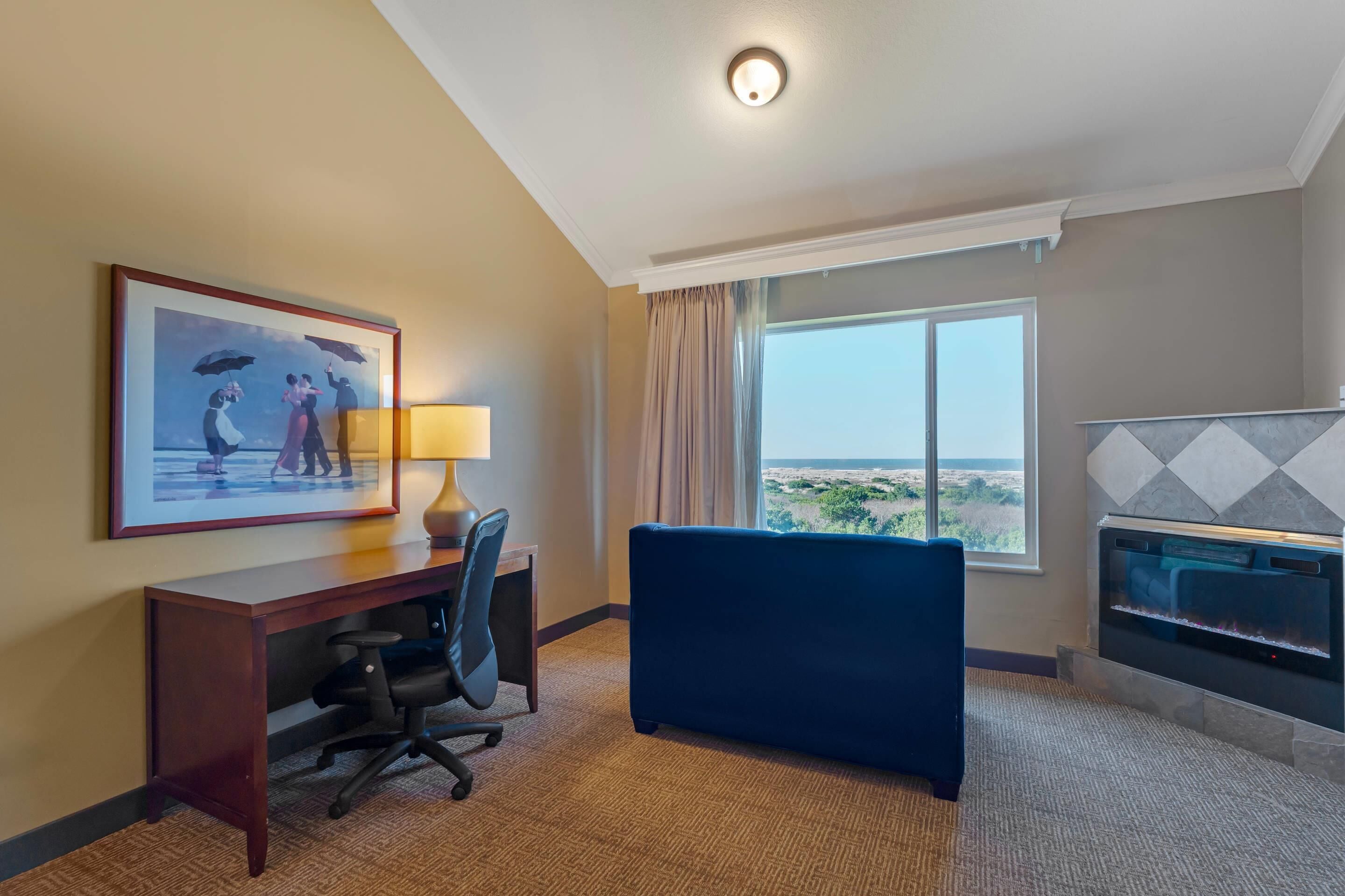 Habitación de hotel con escritorio, silla, un cuadro, sofá, chimenea y un ventanal con vistas.