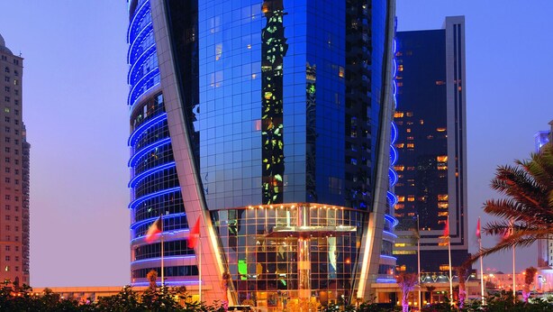 A modern, curved glass building with a prominent entrance and a flagpole. - Qabila Westbay Hotel by Marriott (Doha)
