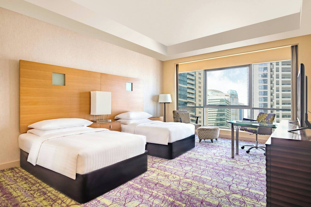 Habitación de hotel con dos camas, un escritorio, una silla y un ventanal con vistas a la ciudad.