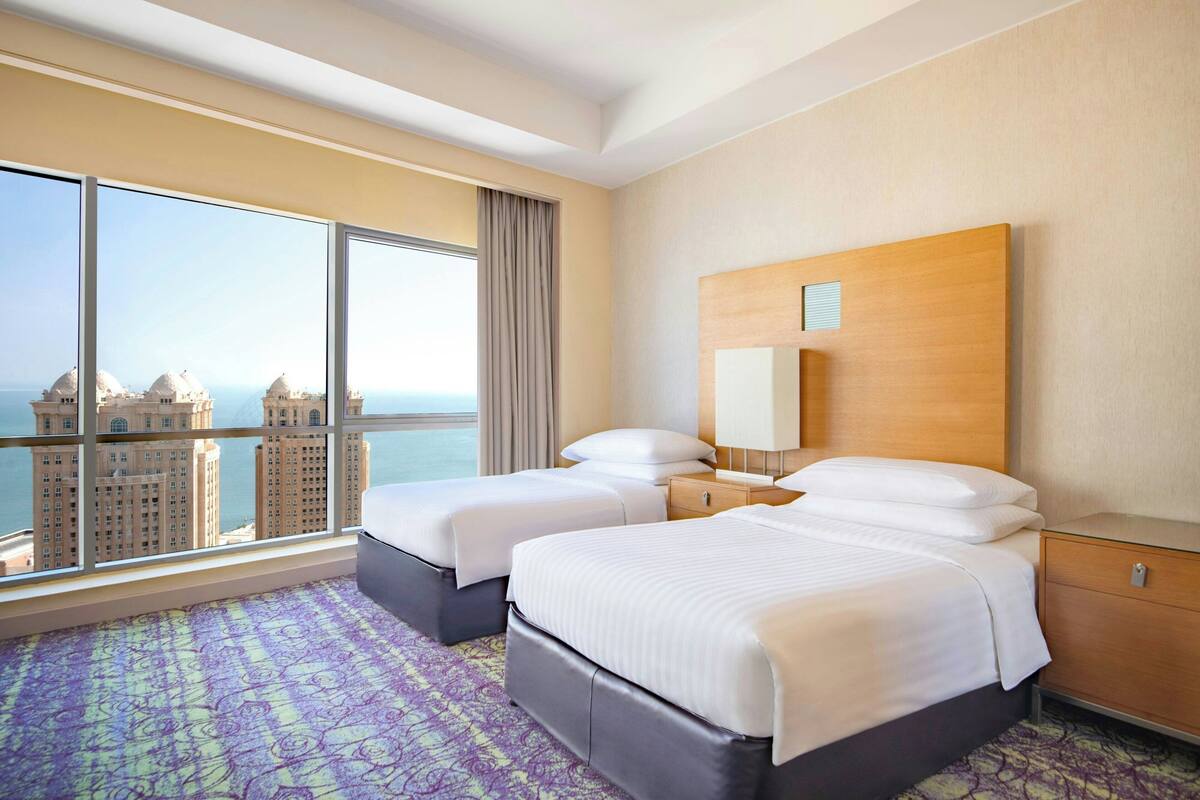 Una habitación de hotel con dos camas, un ventanal con vistas a la ciudad, un cabecero de madera y una alfombra estampada.