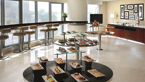 A modern hotel lobby with a central buffet table displaying various pastries and desserts, surrounded by seating areas and large windows offering city views. - Qabila Westbay Hotel by Marriott (Doha)