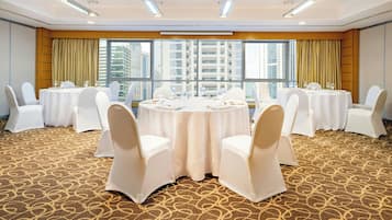 Un salón de conferencias con mesas redondas y sillas blancas, ventanas amplias con cortinas y una alfombra estampada.
