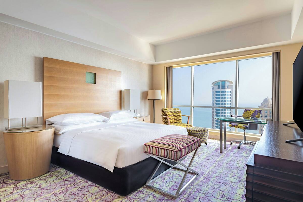 Habitación de hotel con cama, mesitas de noche, escritorio, silla y un amplio ventanal con vista a la ciudad.